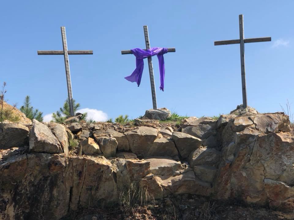 Three wooden crosses on a rocky hillside against a blue sky, the centre cross draped with a purple cloth
