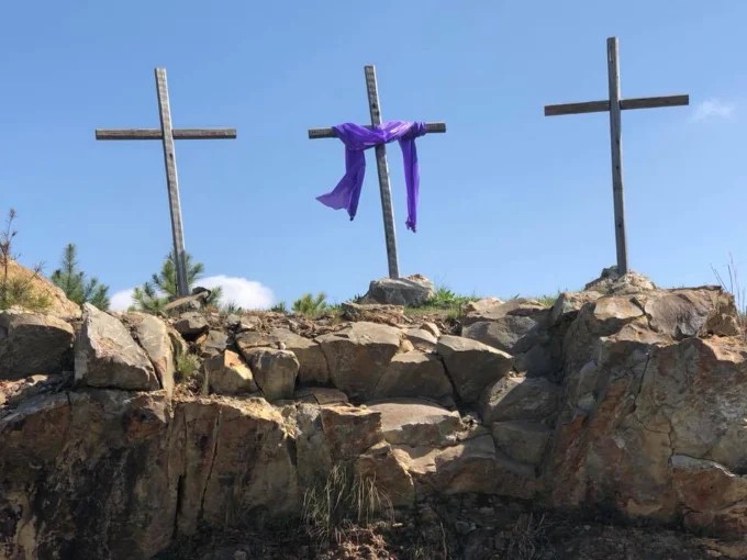 Three wooden crosses on a rocky hillside against a blue sky, the centre
                    cross draped with a purple cloth