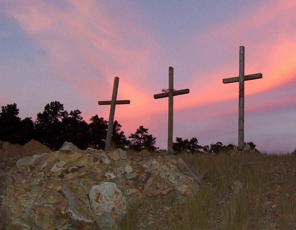 Three wooden crosses on a rocky hillside silhouetted against a pink and
                    purple sunset sky